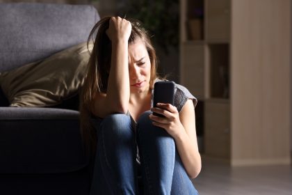 Front view of a sad teen checking phone sitting on the floor in the living room at home with a dark background